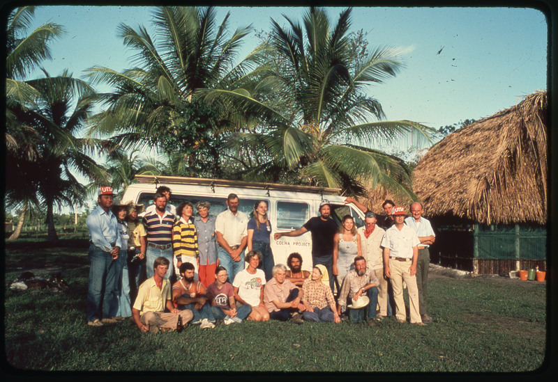 A photographic slide of a group of people standing together in front of a van. There are palm trees in the background and a palm leaf roofed house.