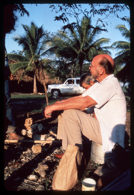 A photographic slide of Donald Crabtree sitting next to another person outside. There looks to be flintknapping materials on the ground.