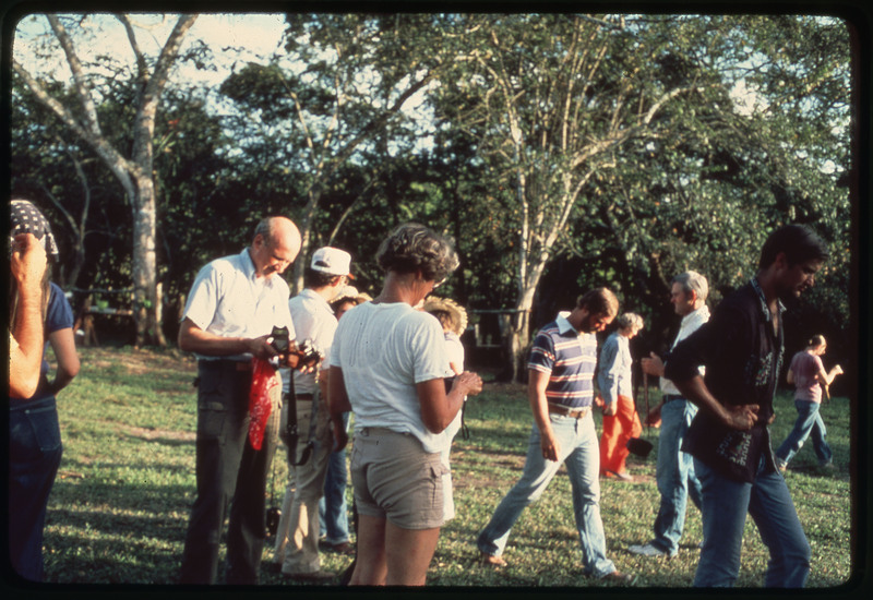 A photographic slide of many people outside talking and holding cameras. There are many trees in the background.