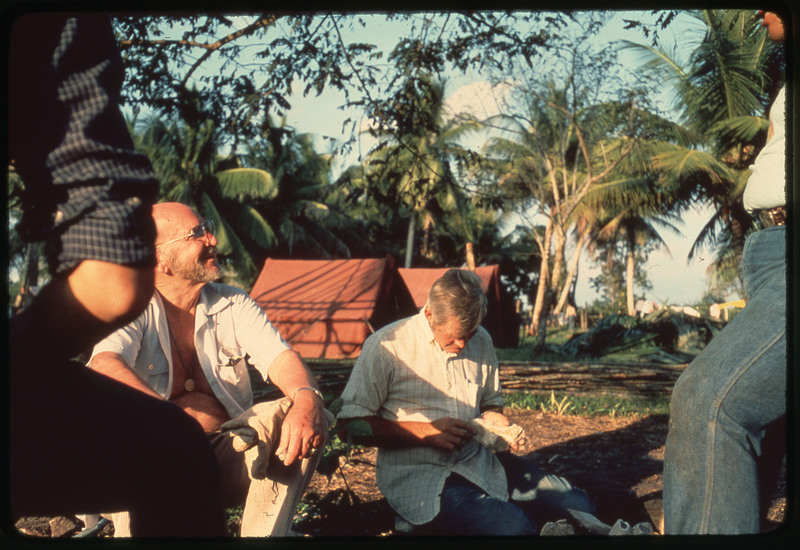 A photographic slide of Donald Crabtree and many other people sitting outside and talking. They appear to be flintknapping. They are in a tropical place.