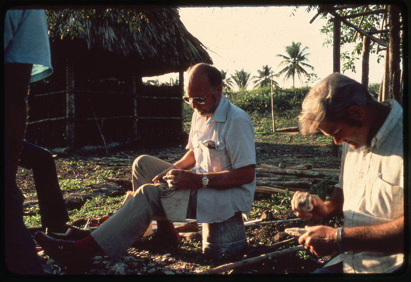 A photographic slide of Donald Crabtree and another person flintknapping. They are in a tropical place with palm trees in the background.