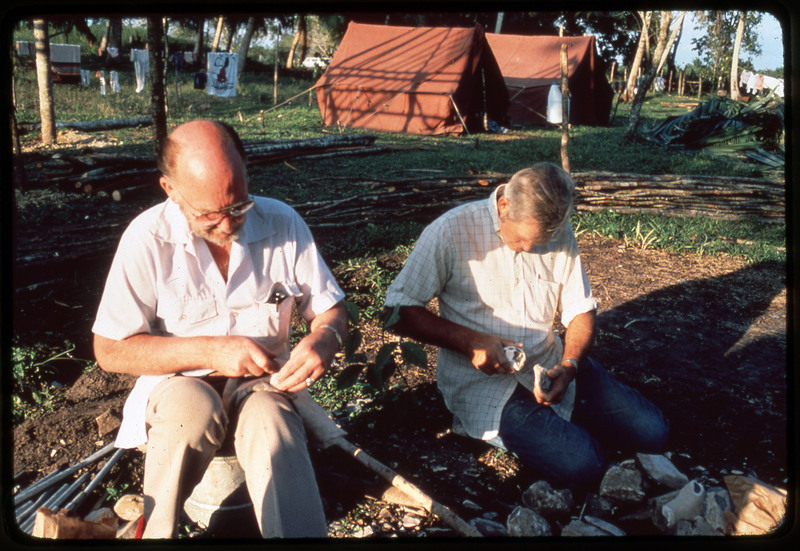 A photographic slide of Donald Crabtree and a friend flintknapping. They are in a tropical place with tents in the background.