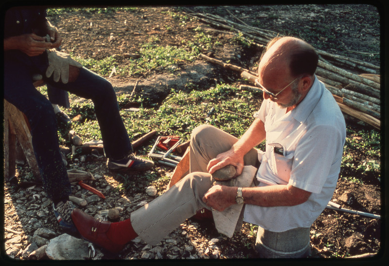 A photographic slide of Donald Crabtree flintknapping using a large stone. They are outside and there are branches in the background.