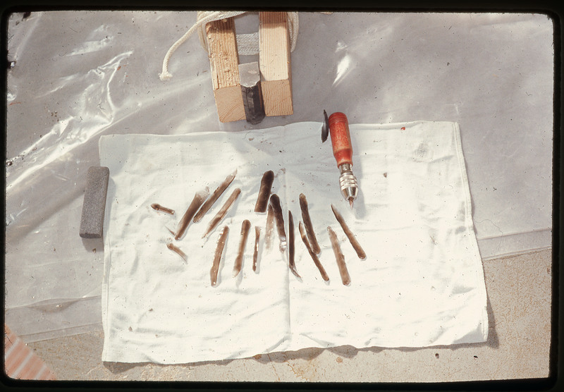 A photographic slide of flaked materials in long vertical shapes. There is a copper tipped tool and chest crutch in the image.