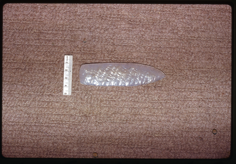 A photographic slide of a grey biface next to a ruler on carpet.