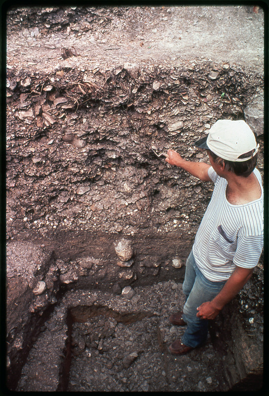 A photographic slide of an archaeological site showing stratigraphy of a pit. There is a man in the pit pointing to something in the stratigraphy.