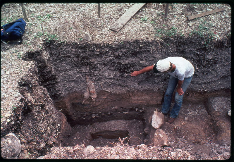 A photographic slide of an archaeological site showing the stratigraphy of the area excavated. There is a man pointing to something on the side of the pit.