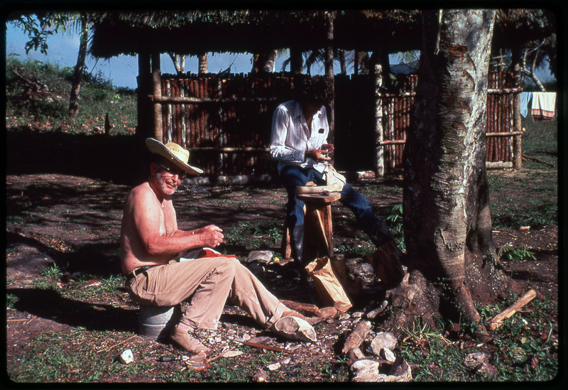 A photographic slide of two people flintknapping next to a palm tree. There is a hut in the background.