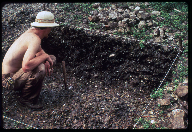 A photographic slide of a man sitting in an archaeological pit. The pit is very rocky and could contain flakes.