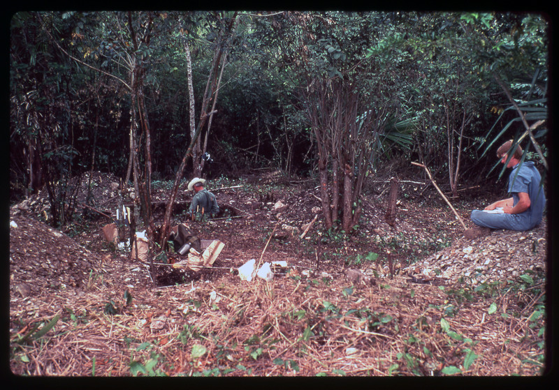 A photographic slide of an archaeological site. There are multiple pits and two people working. They seem to be in a tropical area