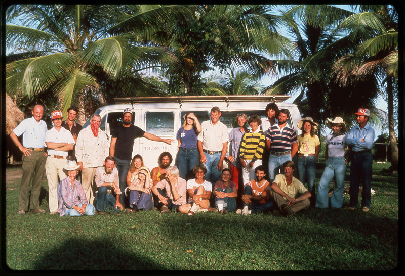 A photographic slide of a large group of people posed together outside a van. There are palm trees in the background. This is a similar photo to ce.81.21.25.