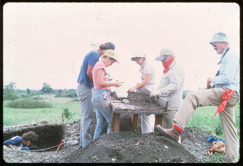 A photographic slide of a group of people working at an archaeological site. There are archaeological pits in the background. The people are sorting through the soil on a table.