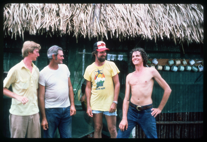 A photographic slide of four men standing outside a straw building. There are mugs in the background and they appear to be in a tropical area.