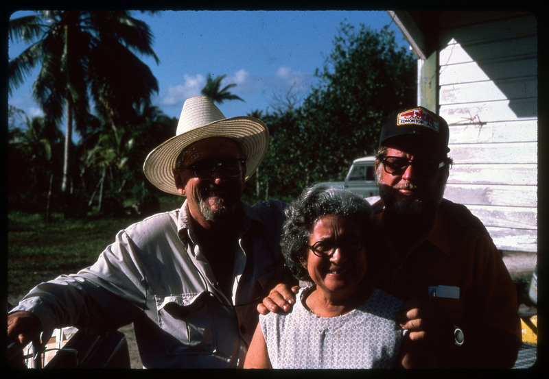 A photographic slide of three people standing together outside. They are posing in a tropical place.