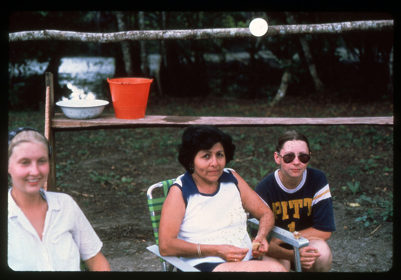 A photographic slide of three people sitting outside together. There is a table behind them and they are eating snacks.