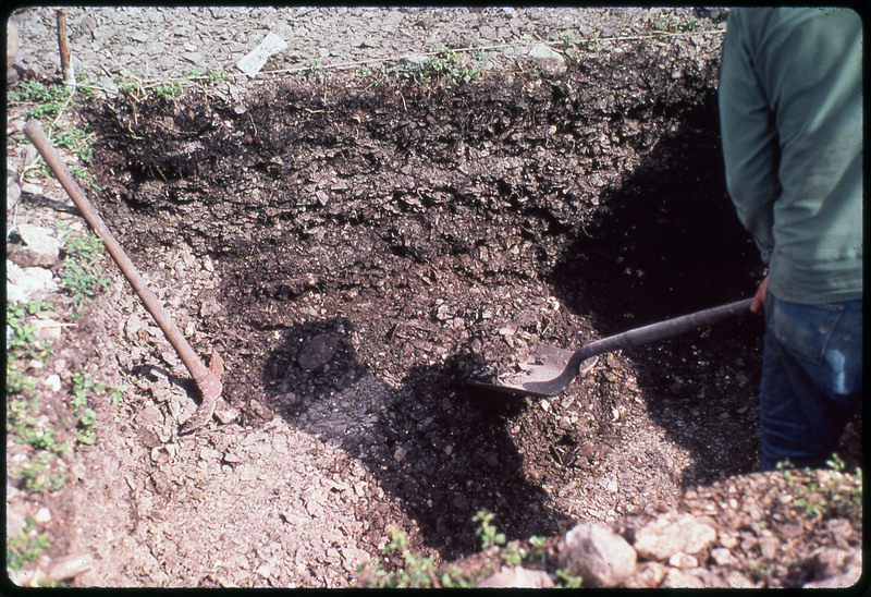 A photographic slide of an archaeological pit. The area is very rocky and there is a person shoveling the pit.