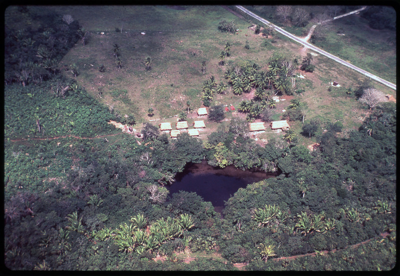 A photographic slide of huts built in a field. It is a tropical area and there seems to be a large pit next to the huts.