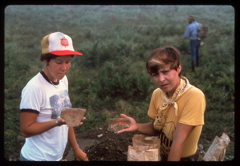 A photographic slide of two people showing pieces of pottery to the camera. There is a field in the background and someone walking.