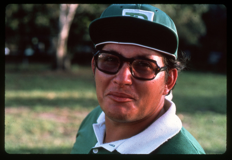 A photographic slide of a man wearing glasses, a green hat, and a green collared shirt. The man is outside.