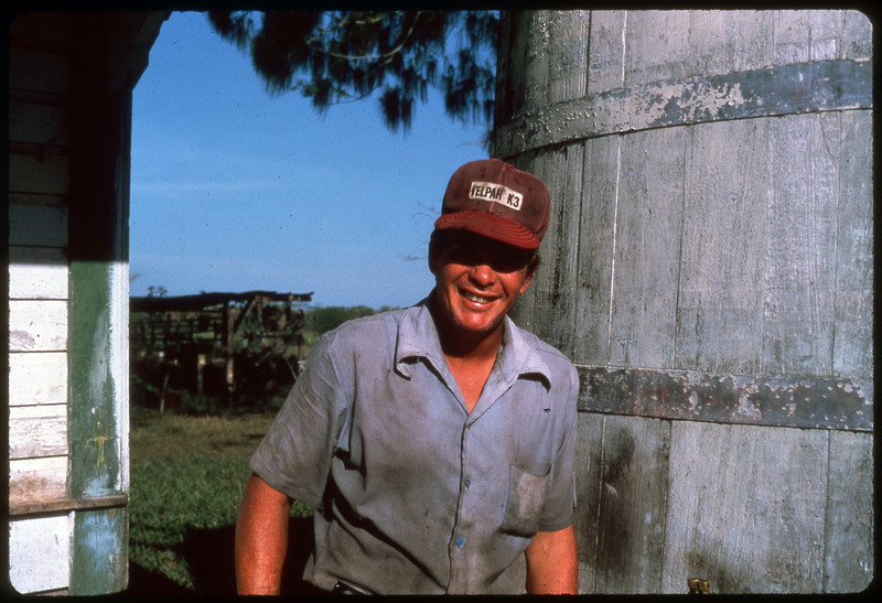 A photographic slide of a man standing outside in work clothes. He is smiling at the camera and there is farm equipment in the background.