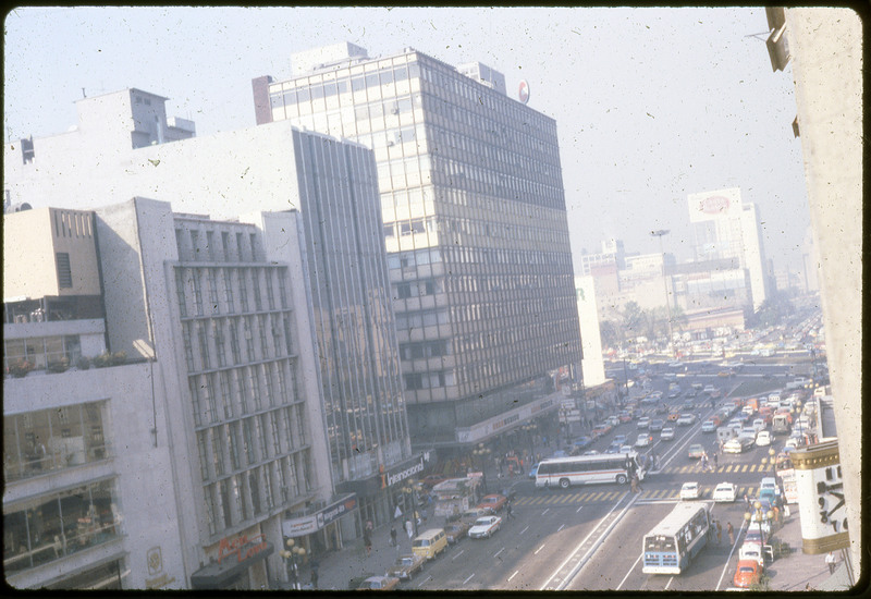 A photographic slide of a city street lined with cars, skyscrapers, and people on the sidewalks.