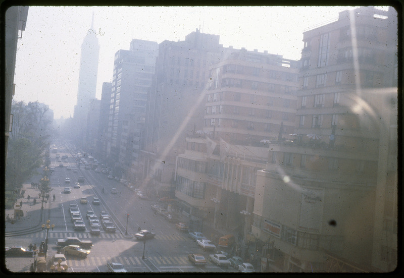 A photographic slide of a city street. There are skyscrapers along the road and many cars on the road. There is a park on one side of the road.