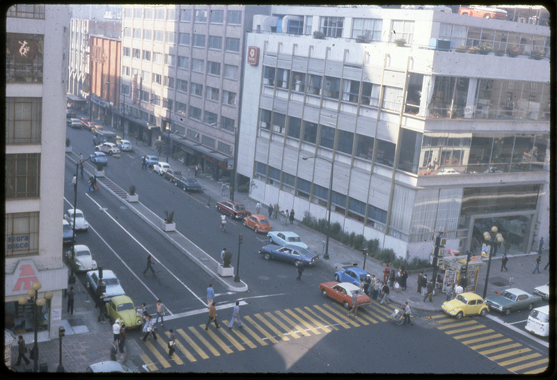 A photographic slide of a city block corner. There are many cars on the road and pedestrians crossing the street. There are many large buildings along the street.