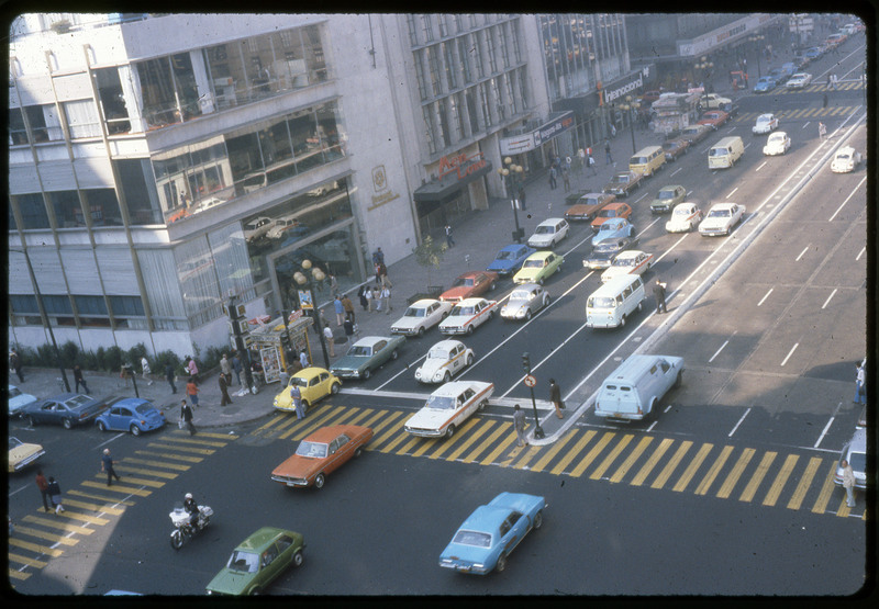 A photographic slide of a city block corner. There are people walking the streets and the sidewalk. Many cars are on the road.