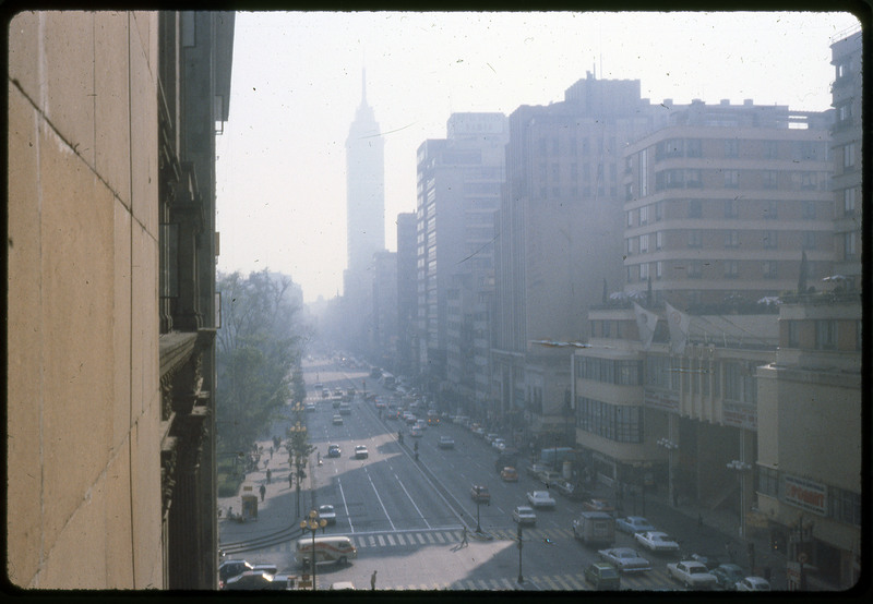 A photographic slide of a city street. Something that resembles the empire state building is in the background. There are many cars on the street and trees on the side.