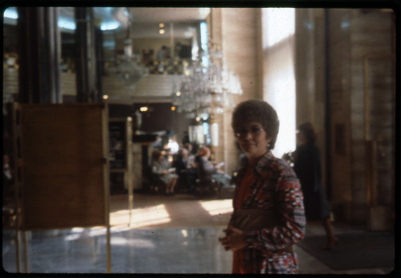 A photographic slide of a woman standing in a nice building. There is a chandelier in the background and people sitting.