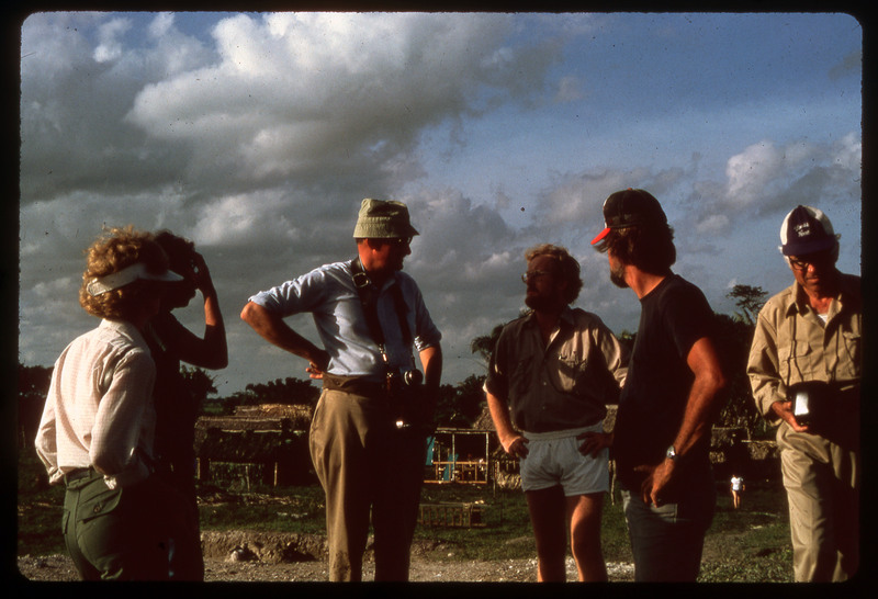 A photographic slide of people standing together talking outside in a tropical area. There are structures in the background.