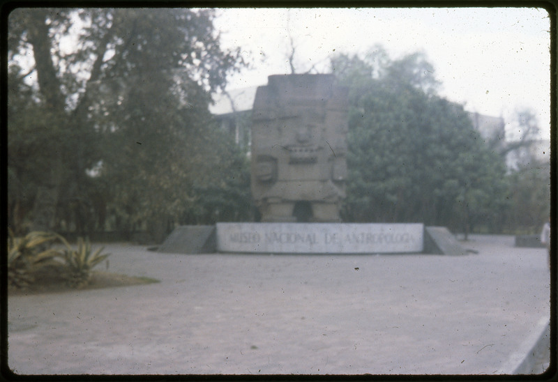 A photographic slide of the entrance to the Museo Nacional de Antropologia. There is a large sculpture outside. This is likely the Museum of Anthropology in Mexico.