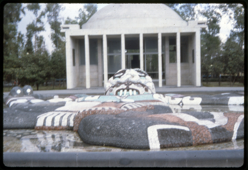 A photographic slide of a colorful sculpture with a face outside. There is a building in the background and trees.