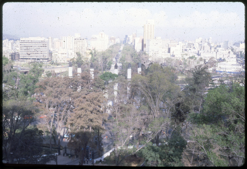 A photographic slide of a cityscape. There are many trees in the foreground. There are artistic pillars pictured and a road in the background.