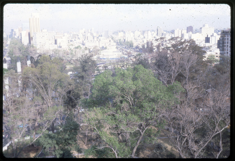 A photographic slide of a tree lined cityscape. There are roads in the background and many skyscrapers.