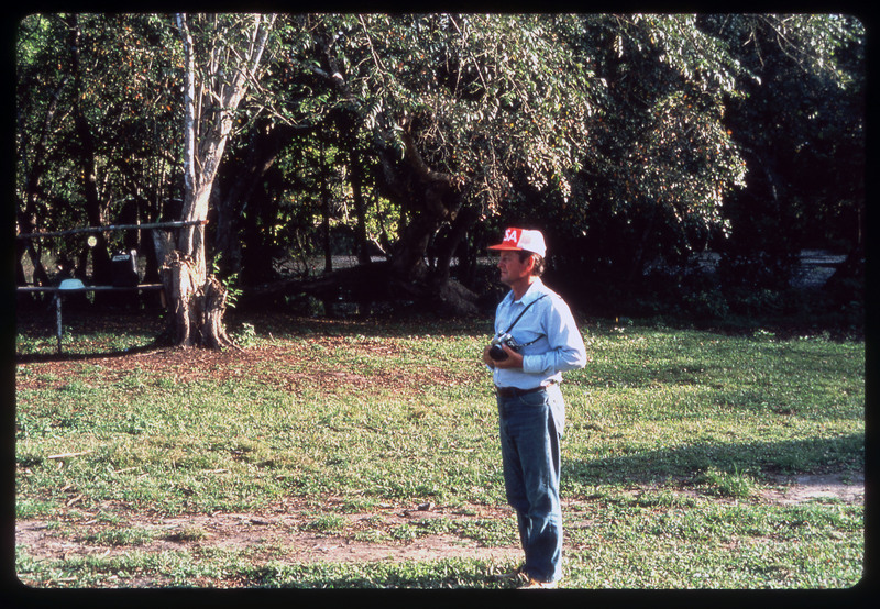 A photographic slide of a man standing outside with a camera. He is in a field with trees behind him.