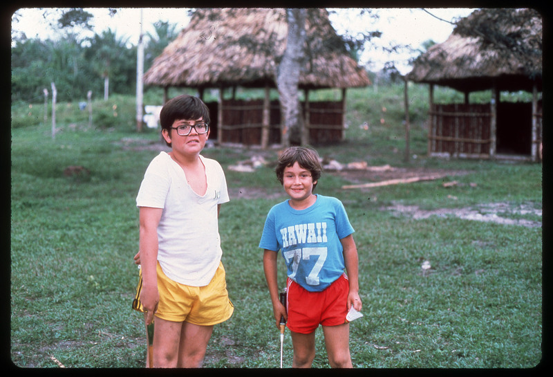 A photographic slide of two young boys standing on a lawn in a tropical area. One boy is wearing a Hawaii shirt. They are holding badminton rackets and a birdie.