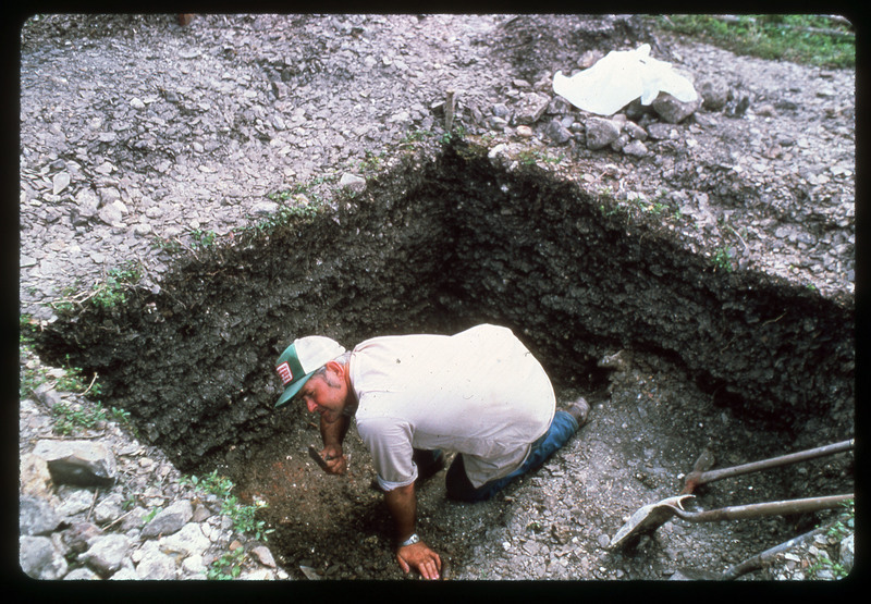 A photographic slide of an archaeological site with a man digging inside. There are many small rocks from the pit, possibly flakes. There are shovels in the pit.