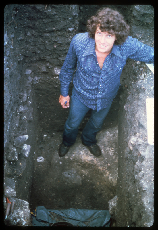 A photographic slide of a man standing in an archaeological pit. The pit shows stratigraphy.