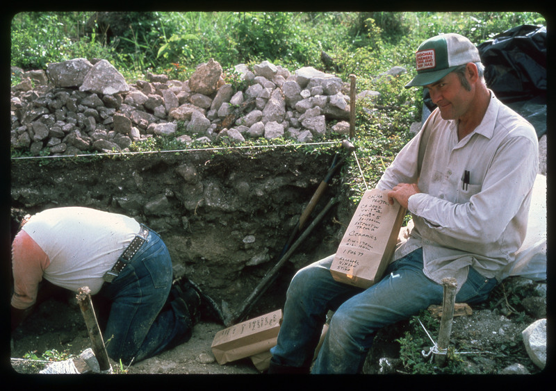 A photographic slide of a man sitting on the edge of an archaeological pit holding a bag. There is another person working in the pit.