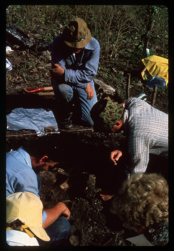 A photographic slide of an archaeological site with people analyzing material in the pit. There are various archaeological tools.