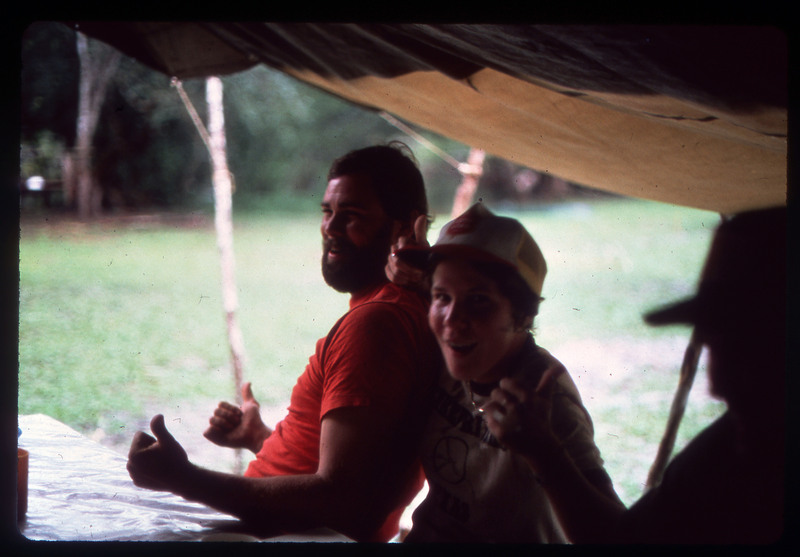 A photographic slide of two people laughing outside under a tent. There is a third person in shadows of the image.