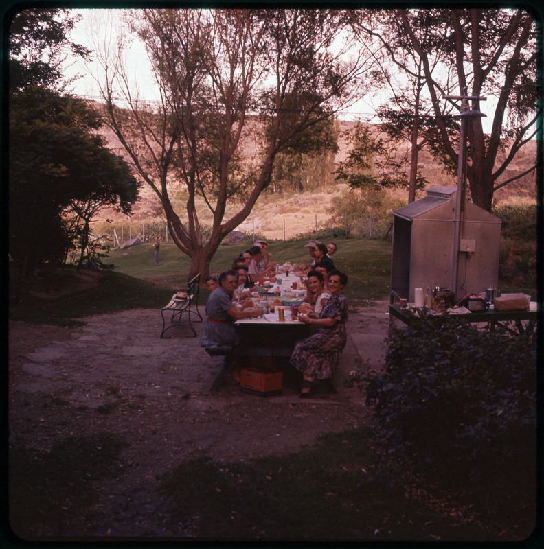 A photographic slide of people sitting together at an outdoor table and eating. There are many people and a big backyard behind them.