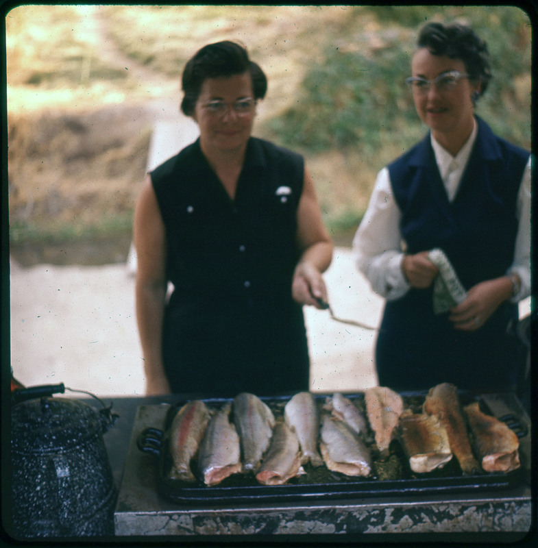 A photographic slide of two women grilling fish outdoors. One woman is holding a spatula.