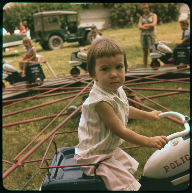 A photographic slide of a young child on a playground merry go round of motorcycles. There are other children in the background.