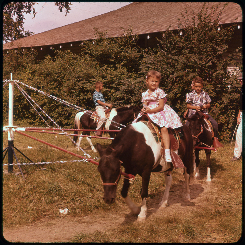 A photographic slide of kids on ponies chained to a spinning piece of equipment. They are outside.