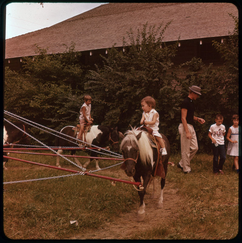 A photographic slide of kids on ponies chained to a spinning piece of equipment. They are outside and there are people not on ponies on the side.