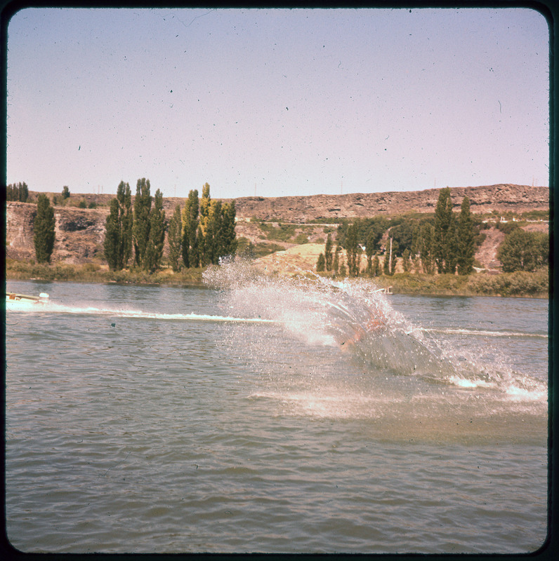 A photographic slide of a person falling in the water on water skis. There is a big splash and trees and hills in the background.