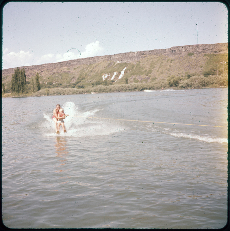 A photographic slide of a man holding a child while water skiing. There are mountains in the background.