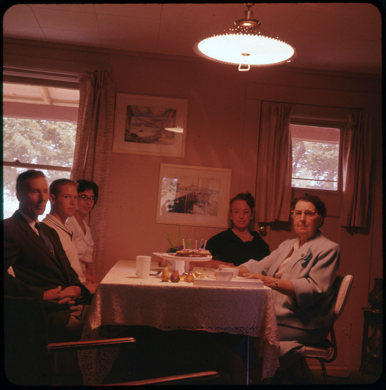 A photographic slide of five people sitting at a table. There is a cake with candles on the table and Evelyn Crabtree is sitting at the table.